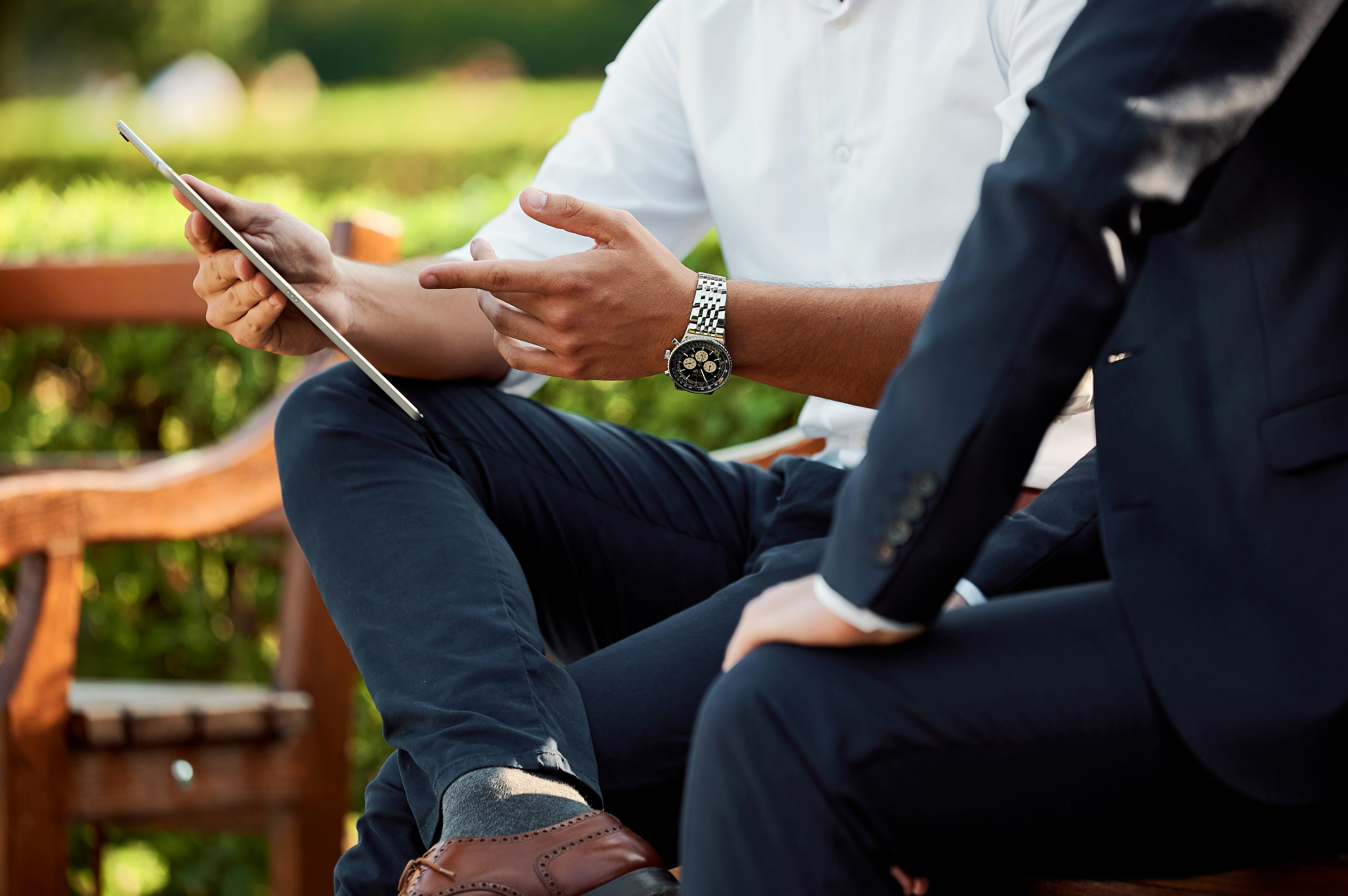 two men sit on a park bench and look at a tablet